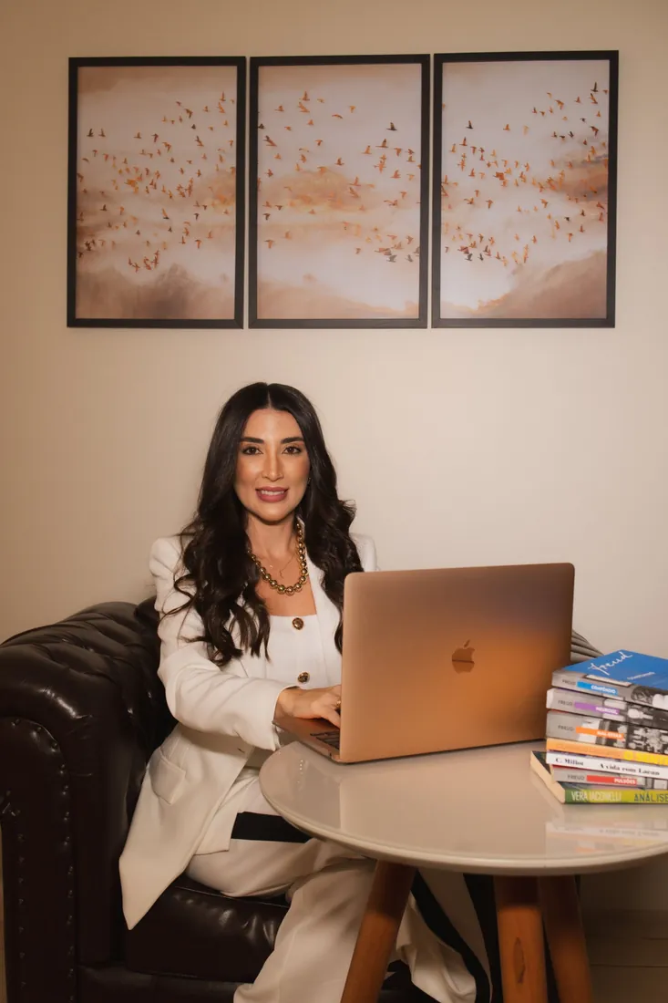Jennifer sorrindo diante do notebook, com livros de psicanálise sobre a mesa