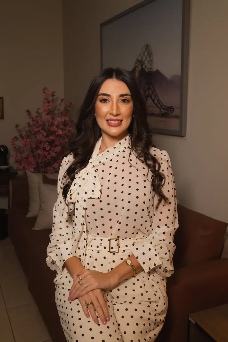 Jennifer sorrindo na sala do consultório, com o sofá, flores e um quadro ao fundo